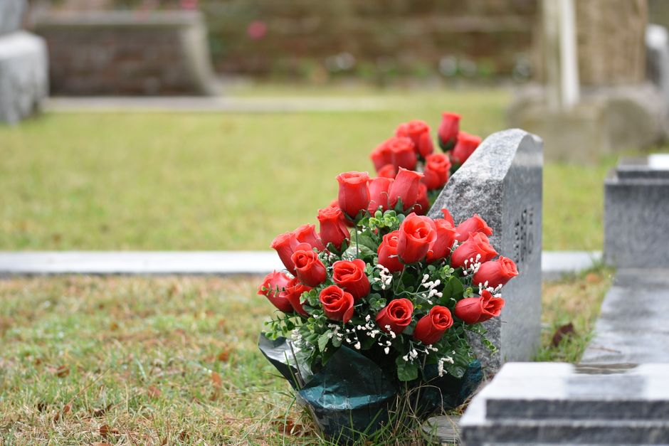 A bouquet of fresh red roses with tightly closed buds and vibrant green leaves, arranged with small white filler flowers and wrapped in dark green tissue paper. The bouquet is placed at the base of a 