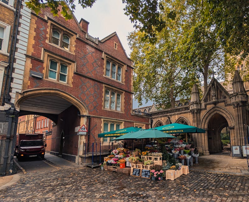 A flower stall situated on a cobbled street beneath an arched brick passageway, displaying a vibrant array of fresh flowers including pink roses, yellow daisies, and purple tulips arranged in wooden c