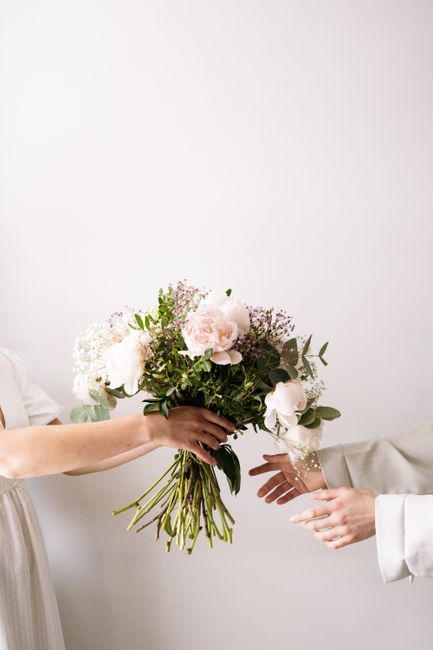 A person hands a large, fresh floral bouquet featuring soft pink peonies, white roses, and delicate sprigs of baby's breath, accented with lush green foliage. The bouquet is arranged in a natural, rou
