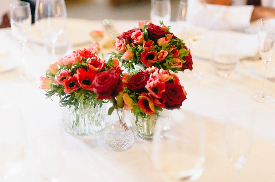 A close-up view of three floral arrangements on a white tablecloth, featuring vibrant red and pink roses, tulips, and anemones with dark centers, all arranged in clear glass vases with intricate patte