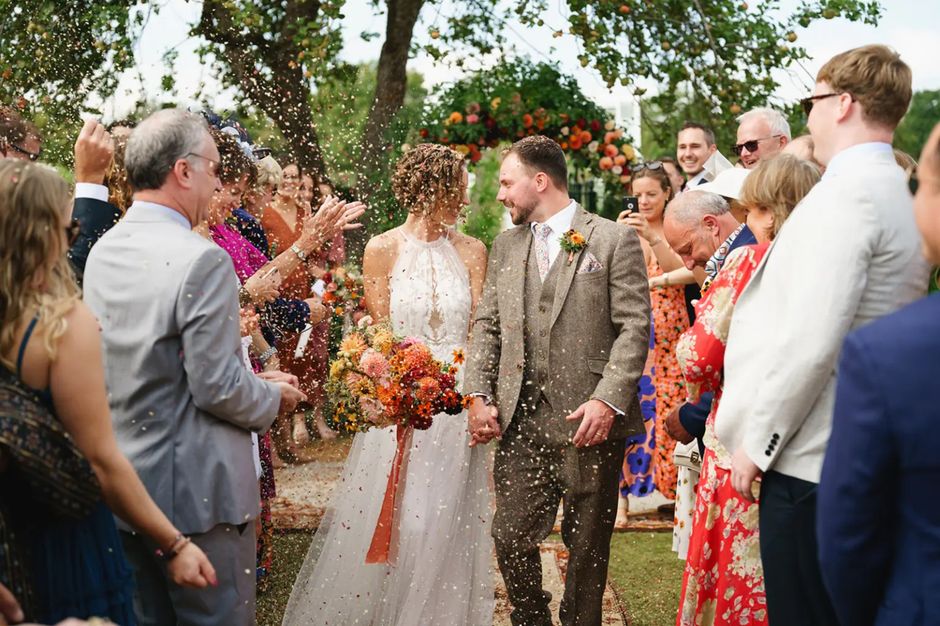 A wedding scene featuring a bride and groom holding hands amidst a jubilant crowd, outdoors in natural daylight with lush green trees in the background. The bride is wearing a sleeveless white lace we