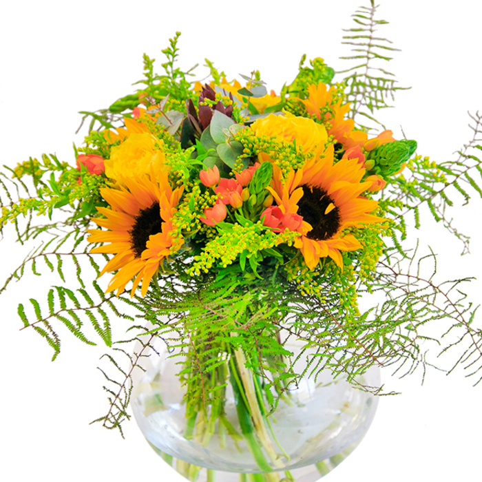 This jubilant bouquet, crafted with the hallmark attention of Flowers Noak Hill, invites the golden charm of the countryside into any room. Its heart features two striking sunflowers, glowing with bright yellow petals and dramatic chocolate-brown centers-much like those that dot the edges of pastureland off Lower Noke Close. Rings of soft yellow filler flowers encircle the sunflowers, lending texture and depth, while scattered coral-orange blooms introduce a friendly warmth reminiscent of early autumn sunsets over Noak Hill's rolling fields. Fern fronds, delicate and lacy, spill outward to soften the silhouette, blending with cool eucalyptus sprigs and crisp, zesty foliage that echo the wild abundance seen in local gardens. The clear, round vase anchors the bouquet, allowing fresh green stems to glimmer through the water-a testament to freshness and expert local floristry. Light bathes every detail, revealing the arrangement's craftsmanship and its balanced, natural grace. Designed for moments of joy, celebration or encouragement, this bouquet makes a fitting centerpiece for gatherings, a cheerful welcome for visitors, or an expressive gift for loved ones anywhere in Noak Hill. The colours, scents, and textures all come together to capture the quiet, uplifting spirit of the area.